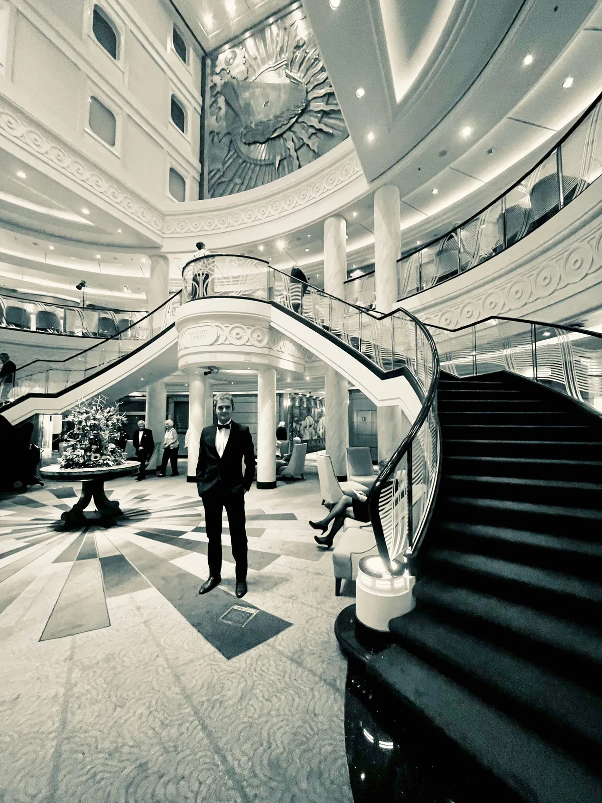 A passenger in a tuxedo standing in the Grand Lobby beneath its sweeping staircase on a gala evening