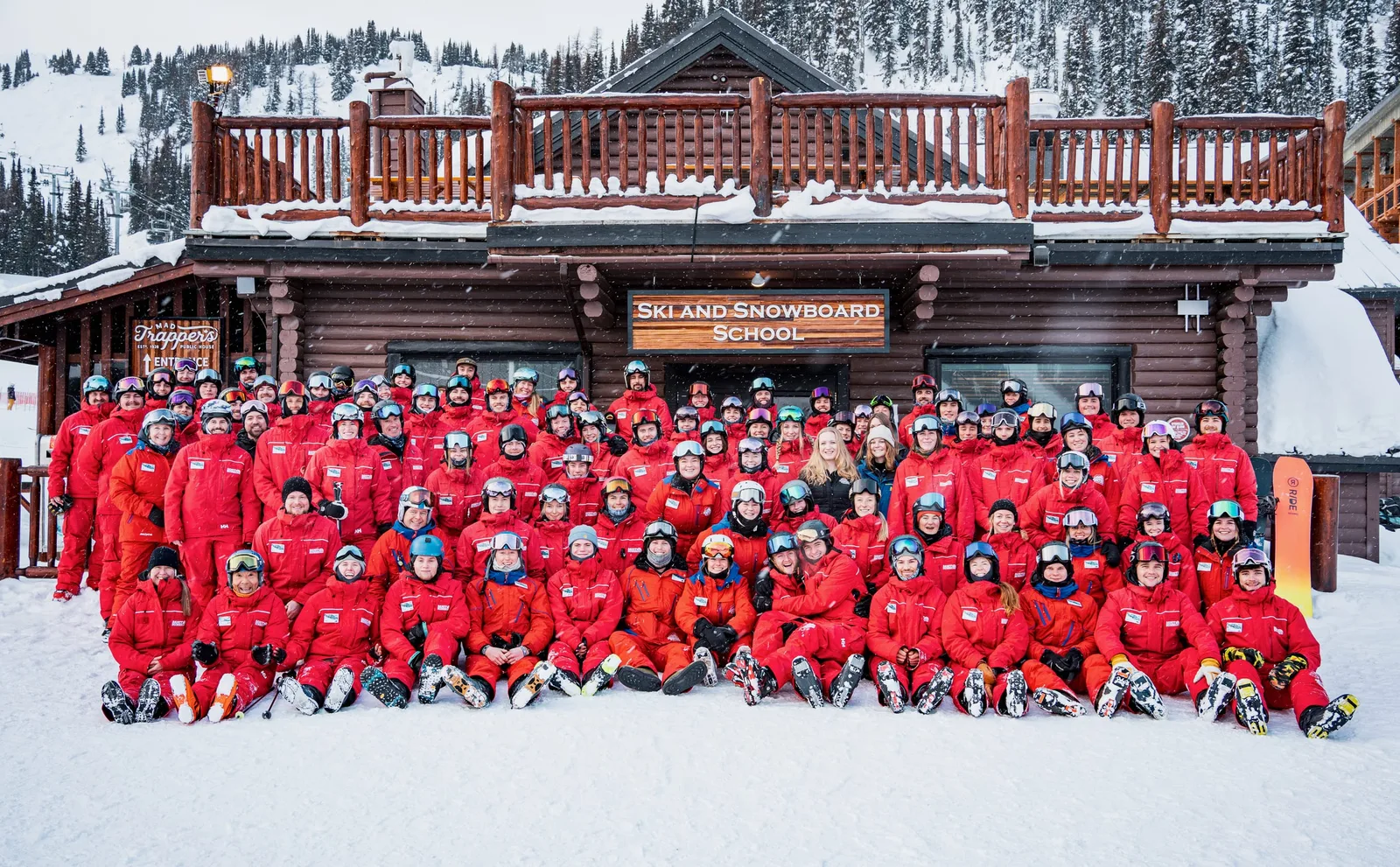 The ski school team in red uniforms gathered in falling snow in front of the wooden Ski and Snowboard School lodge