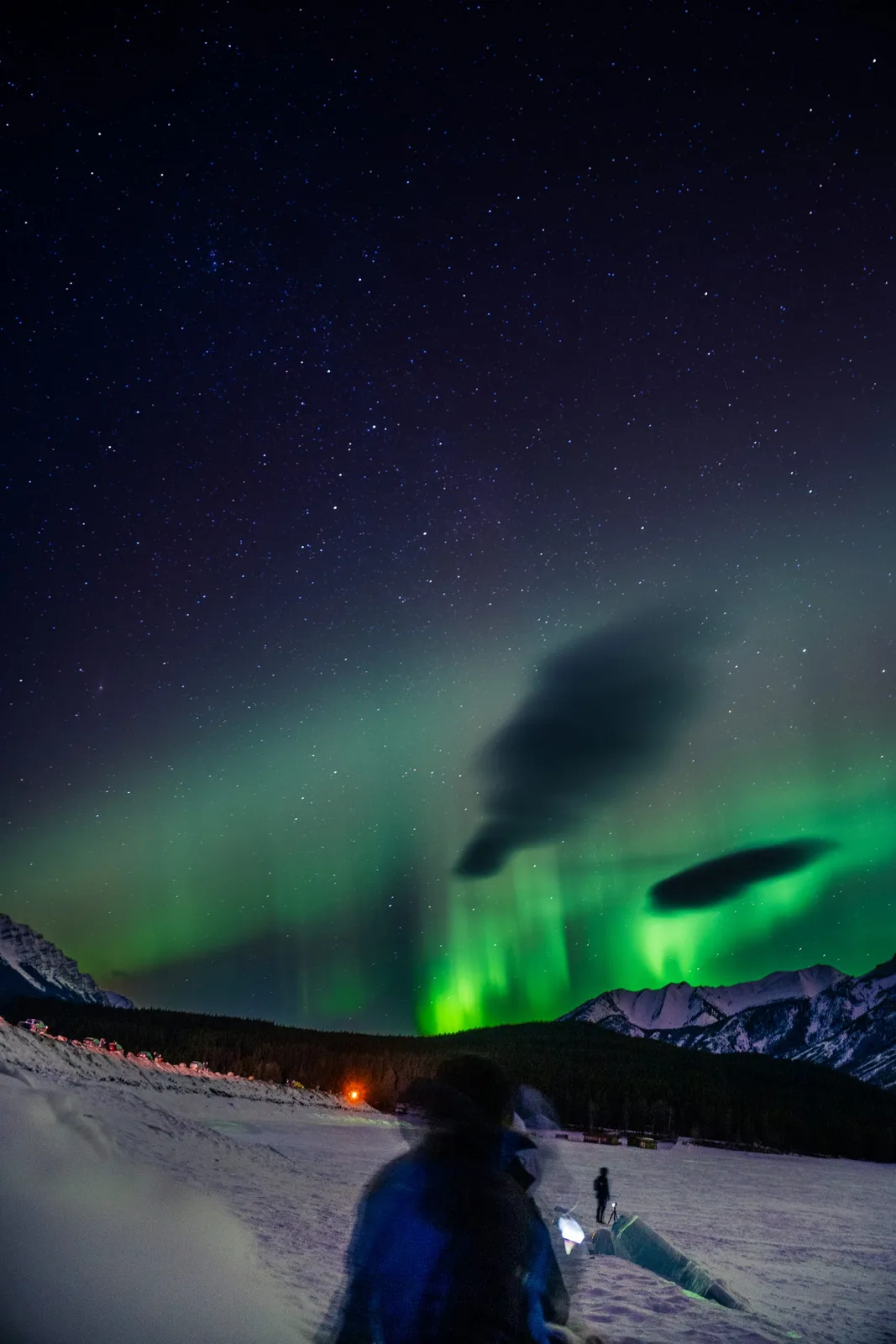 Green northern lights dancing over the Rockies while spectators watch from a frozen lake